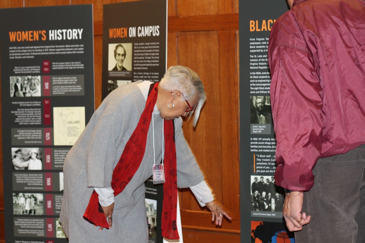 A woman wearing a red scarf leans over to point out a picture on a sign to a man who is partially out of frame. Behind the woman are two additional signs that read Women's History and Women on Campus.