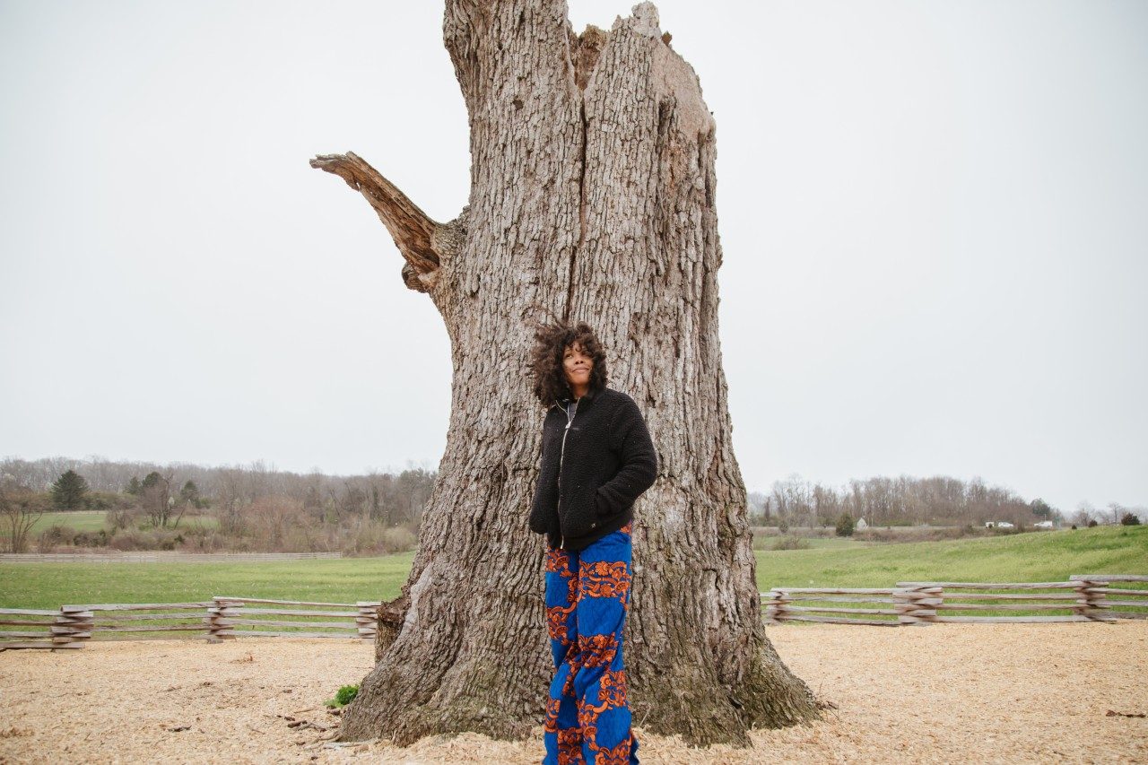 Dr. Kerri Moseley Hobbs stands in of a tall tree trunk. In the background is a fence and field. 